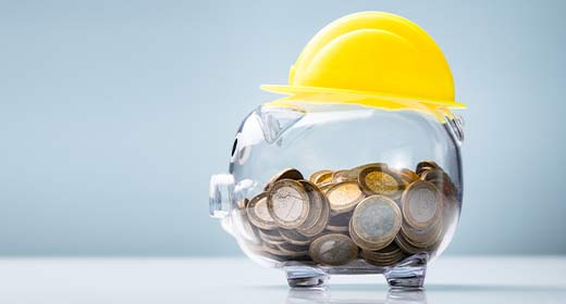 Ein gläsernes Sparschwein mit Münzen und einem Mini-Baustellenhelm in Gelb