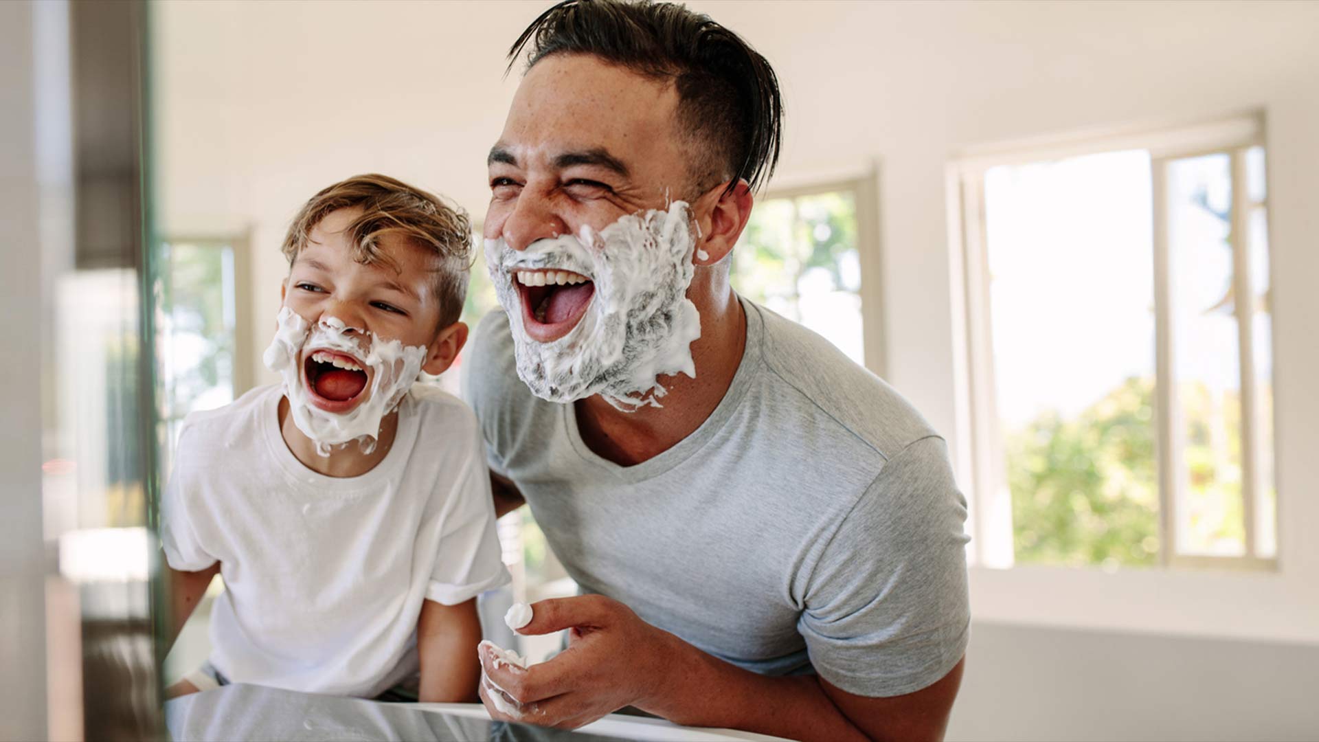 Vater und Sohn mit Rasierschaum im Gesicht lachen in einen Spiegel